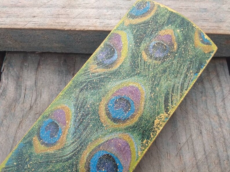 May include: A rectangular wooden bookmark with a peacock feather design. The bookmark is painted in shades of green, blue, purple, and gold. The gold paint is glittery.