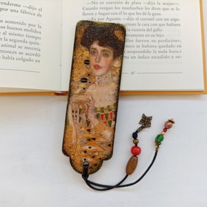 May include: A bookmark with a gold and brown background featuring a painting of a woman with long, flowing hair. The bookmark has a black tassel and a small charm with beads.