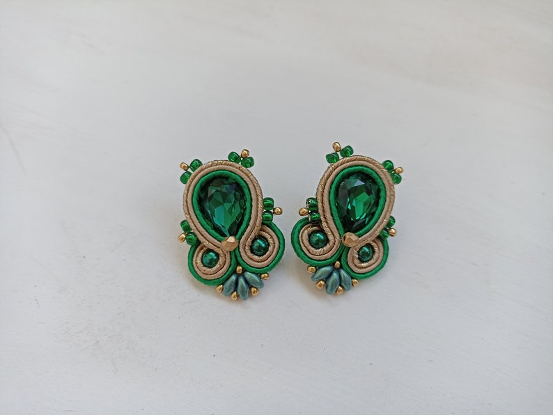 May include: A pair of green and gold earrings with a teardrop-shaped green gemstone in the center of each earring.