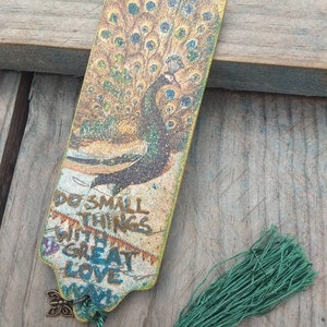 May include: A wooden bookmark with a peacock design and a green tassel. The bookmark has the text "Do small things with great love" printed on it.