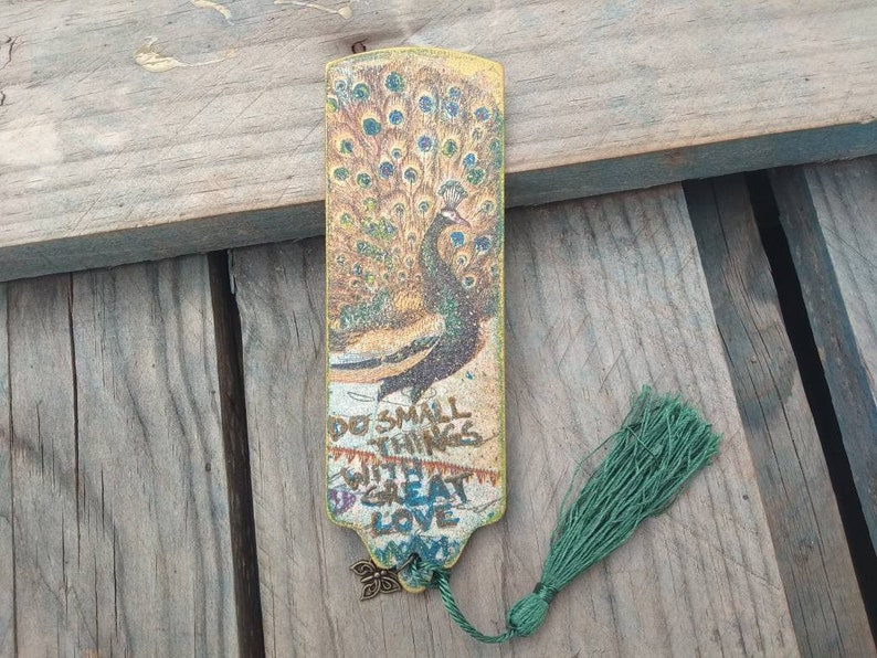 May include: A yellow and green peacock bookmark with a tassel. The bookmark has the text "Do small things with great love" on it.