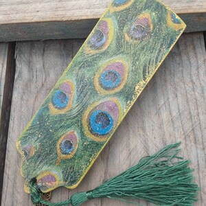 May include: A green bookmark with a peacock feather design and gold accents. The bookmark has a green tassel.
