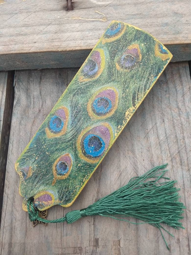 May include: A green bookmark with a peacock feather design and gold accents. The bookmark has a green tassel.