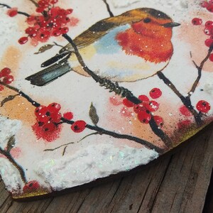 May include: A heart-shaped wooden ornament with a painted image of a robin perched on a branch with red berries. The ornament is covered in white glitter and has a rustic, vintage look.