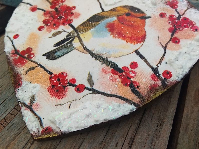 May include: A heart-shaped wooden ornament with a painted image of a robin perched on a branch with red berries. The ornament is covered in white glitter and has a rustic, vintage look.