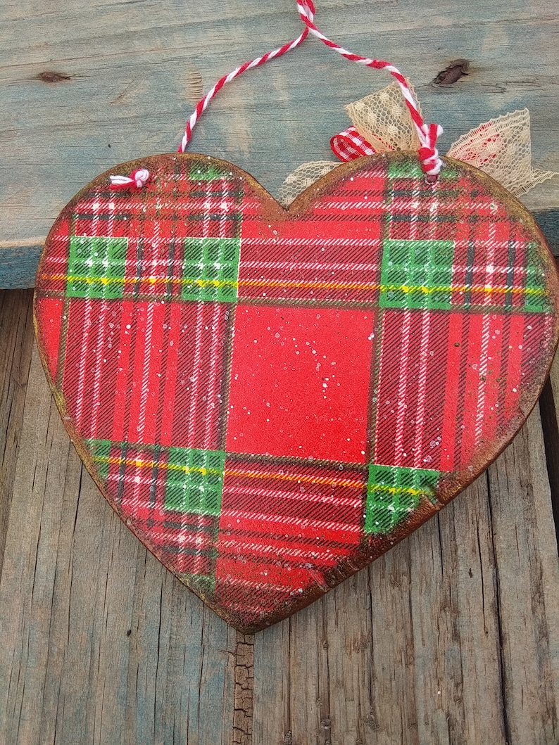 May include: Red, green, and white plaid heart-shaped wooden ornament with a white and red twine hanger and a beige lace bow.