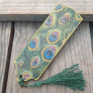 May include: A green and gold bookmark with a peacock feather design. The bookmark has a green tassel.