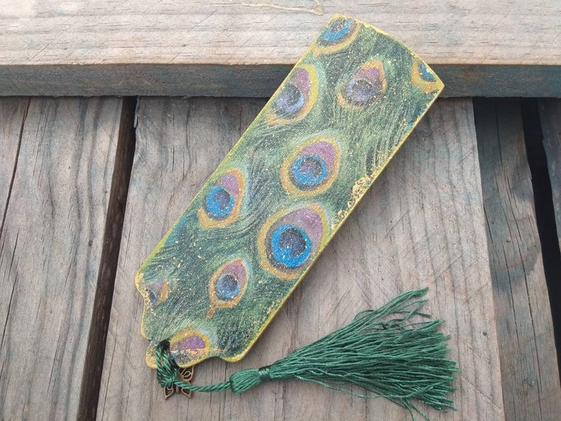 May include: A green and gold bookmark with a peacock feather design. The bookmark has a green tassel.