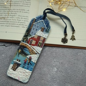 May include: A wooden bookmark with a winter scene painted on it. The bookmark features a small town with snow-covered houses, a bridge, and a couple ice skating on a frozen lake. The bookmark is attached to a blue ribbon with two metal charms, a snowflake and a Christmas tree.