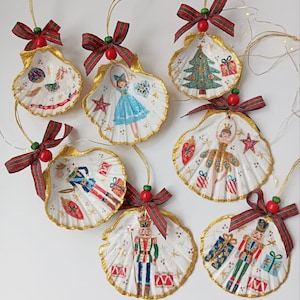 Op de afbeelding: Set van acht handgeschilderde schelp kerstversieringen. Elke versiering heeft een ander feestelijk ontwerp, waaronder een ballerina, een notenkraker, een kerstboom en een meisje dat een hart vasthoudt. De versieringen zijn versierd met gouden glitter en rood-groen geruit lint.