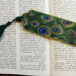 May include: A green and gold bookmark with a peacock feather design. The bookmark has a tassel on the top.