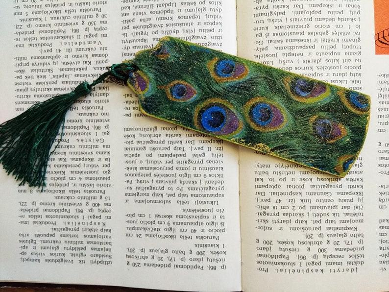 May include: A green and gold bookmark with a peacock feather design. The bookmark has a tassel on the top.