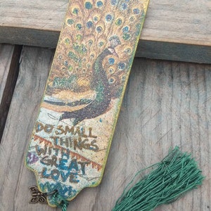 May include: A wooden bookmark with a peacock design and a green tassel. The bookmark has the text "Do small things with great love" printed on it.