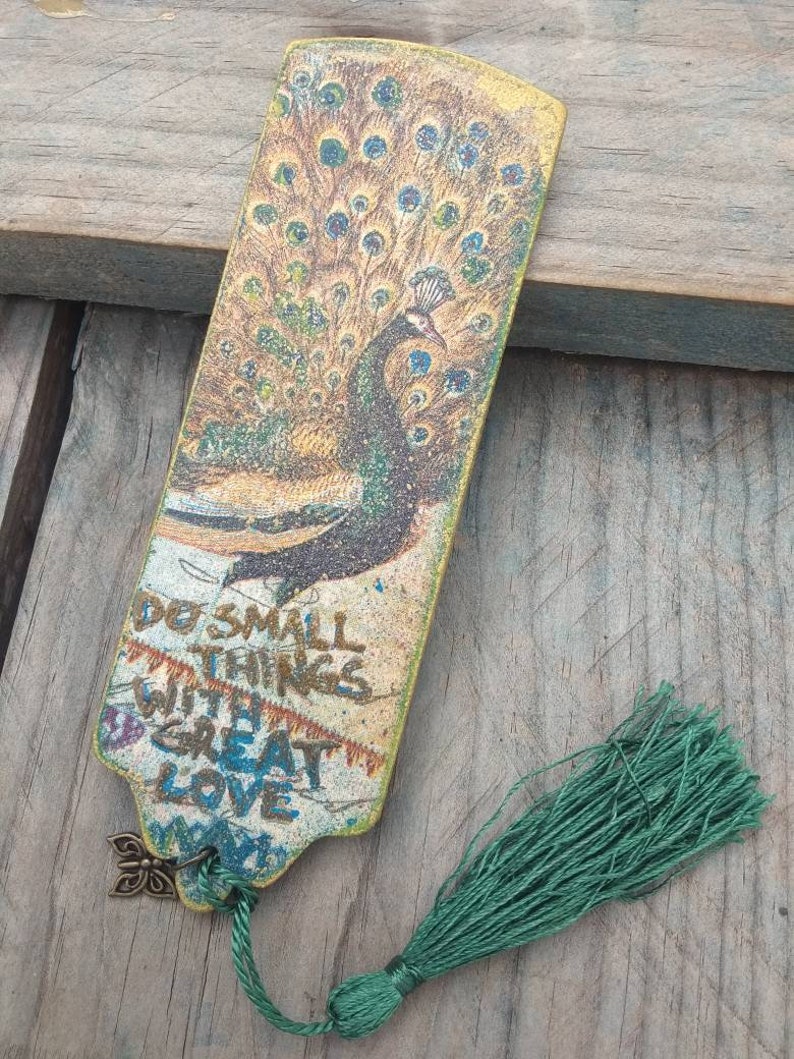 May include: A wooden bookmark with a peacock design and a green tassel. The bookmark has the text "Do small things with great love" printed on it.