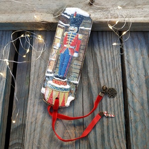 May include: A red and blue Christmas ornament featuring a nutcracker soldier standing on a drum. The ornament has a red ribbon and two metal charms, one with a snowflake and the other with a candy cane.