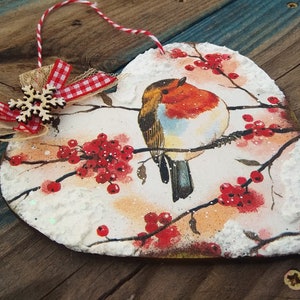 May include: A heart-shaped wooden ornament with a robin perched on a branch with red berries. The ornament is decorated with white snow and glitter. A red and white checkered ribbon with a wooden snowflake is attached to the top.