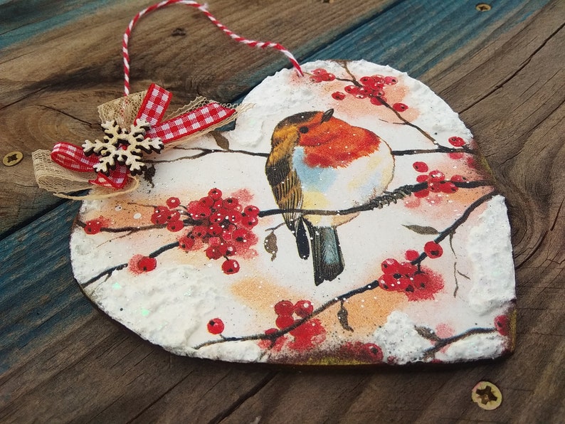 May include: A heart-shaped wooden ornament with a robin perched on a branch with red berries. The ornament is decorated with white snow and glitter. A red and white checkered ribbon with a wooden snowflake is attached to the top.
