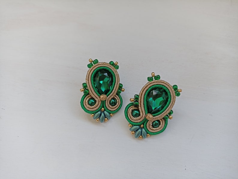 May include: A pair of green and gold earrings with a teardrop-shaped green gemstone in the center of each earring.