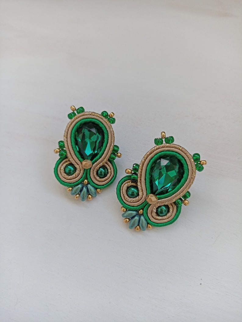 May include: A pair of green and gold earrings with a teardrop-shaped green crystal in the center of each earring.