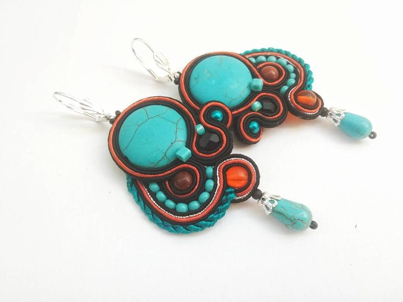 May include: Turquoise and orange statement earrings with a large turquoise stone and smaller turquoise beads. The earrings are made with black and orange thread and have silver hooks.