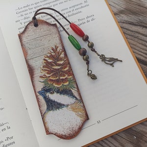 Può includere: Un segnalibro in legno con un'immagine dipinta di un uccello posato su una pigna. Il segnalibro è decorato in tonalità di marrone, bianco e verde. Ha un fiocco con perline rosse, verdi e marroni e un piccolo ciondolo a forma di uccello.