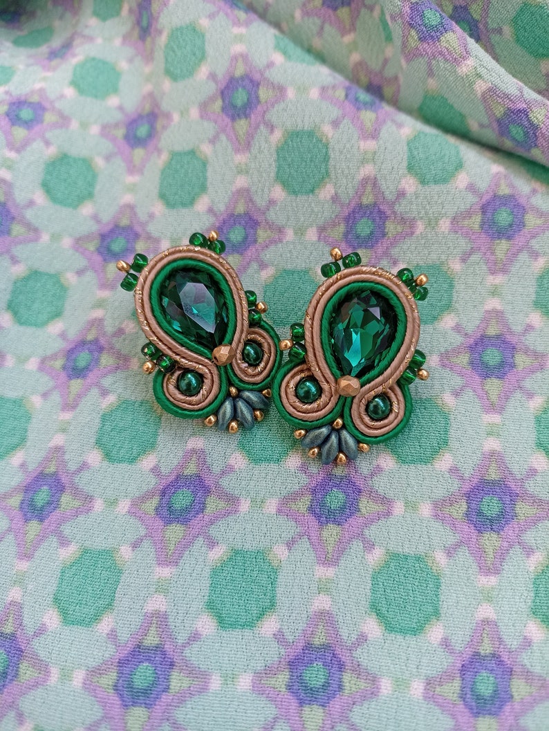 May include: A pair of green and gold earrings with a teardrop-shaped green crystal in the center of each earring.