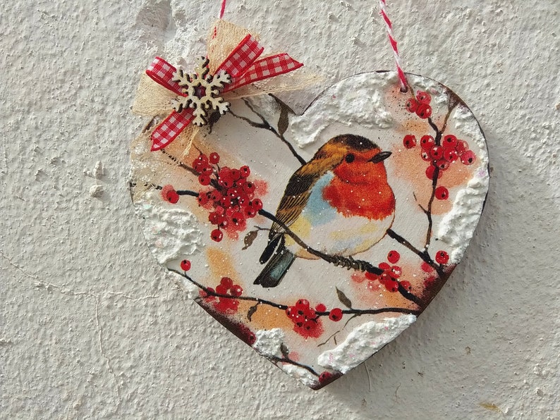 May include: A heart-shaped wooden ornament with a robin perched on a branch with red berries. The ornament is decorated with faux snow and a red and white gingham bow with a wooden snowflake.