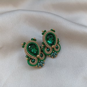 May include: A pair of green and gold earrings with a teardrop-shaped green crystal in the center of each earring.