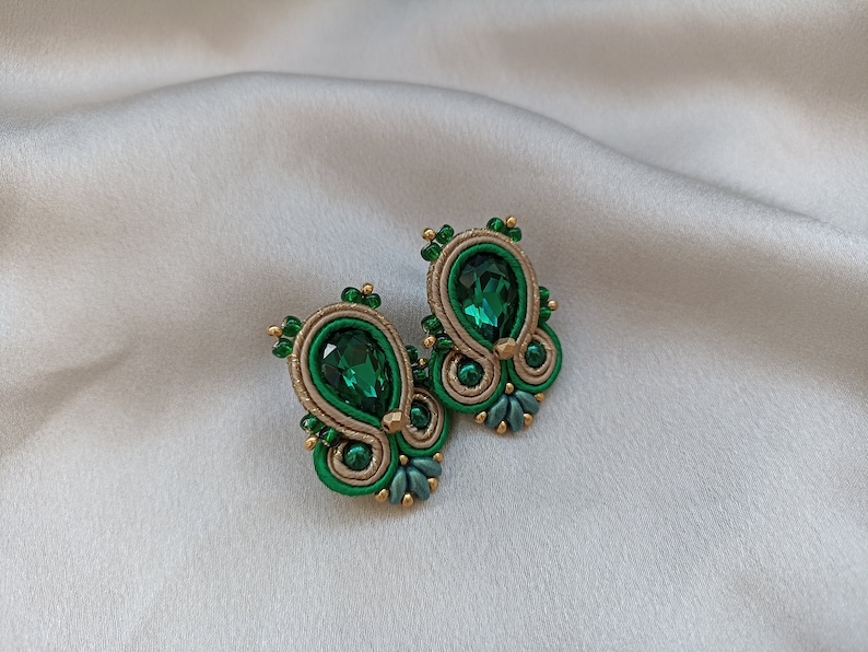 May include: A pair of green and gold earrings with a teardrop-shaped green crystal in the center of each earring.