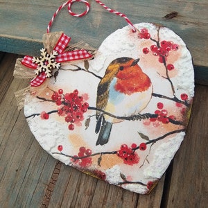 May include: A heart-shaped wooden ornament with a robin perched on a branch with red berries. The ornament is decorated with white snow-like accents and a red and white gingham bow with a snowflake.