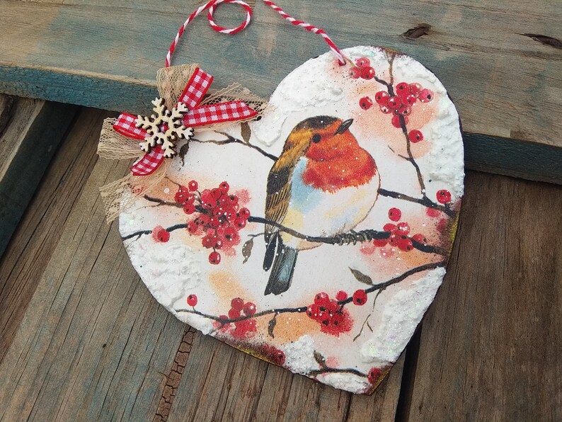 May include: A heart-shaped wooden ornament with a robin perched on a branch with red berries. The ornament is decorated with white snow-like accents and a red and white gingham bow with a snowflake.