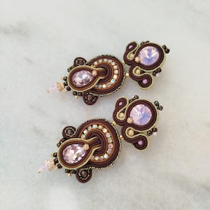 May include: A pair of handcrafted earrings featuring a central oval-shaped pink gemstone, surrounded by intricate brown and gold threadwork. Additional pink and gold accents and small crystals complete the design. The earrings are set against a white marble surface.