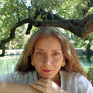 May include: A woman with long brown hair wearing gold earrings with a floral design. She is wearing a white shirt and is sitting with her arms crossed in front of her. The background is a pond with trees and greenery.