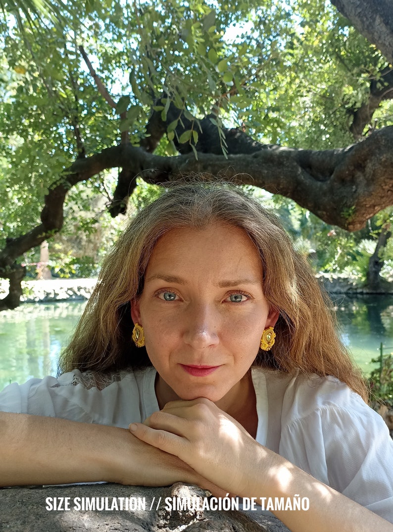 May include: A woman with long brown hair wearing gold earrings with a floral design. She is wearing a white shirt and is sitting with her arms crossed in front of her. The background is a pond with trees and greenery.