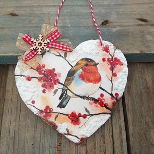 May include: A heart-shaped wooden ornament with a painted image of a robin perched on a branch with red berries. The ornament is decorated with a white snowflake and a red and white gingham bow.