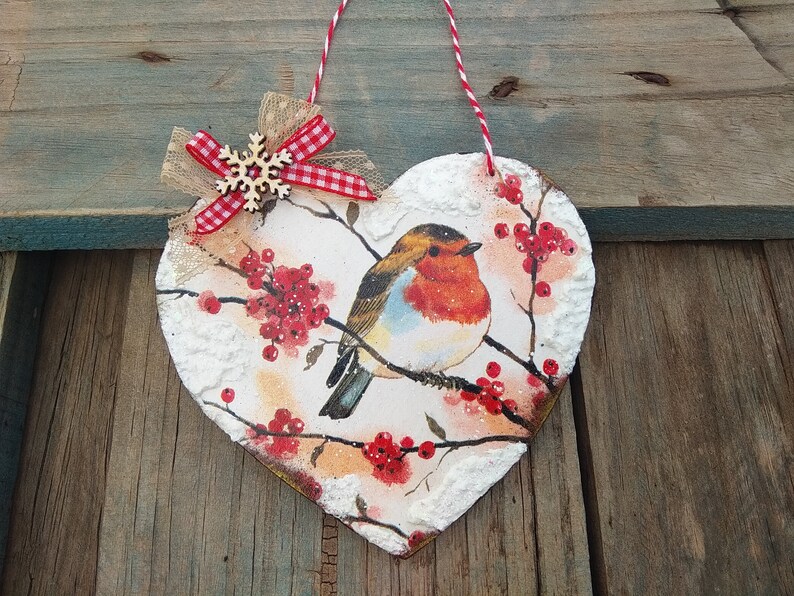 May include: A heart-shaped wooden ornament with a painted image of a robin perched on a branch with red berries. The ornament is decorated with a white snowflake and a red and white gingham bow.
