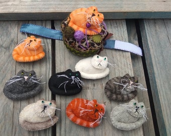 May include: A collection of nine felt cat pincushions in various colours and patterns, including orange tabby, grey tabby, black, and white. The pincushions are arranged on a wooden surface.