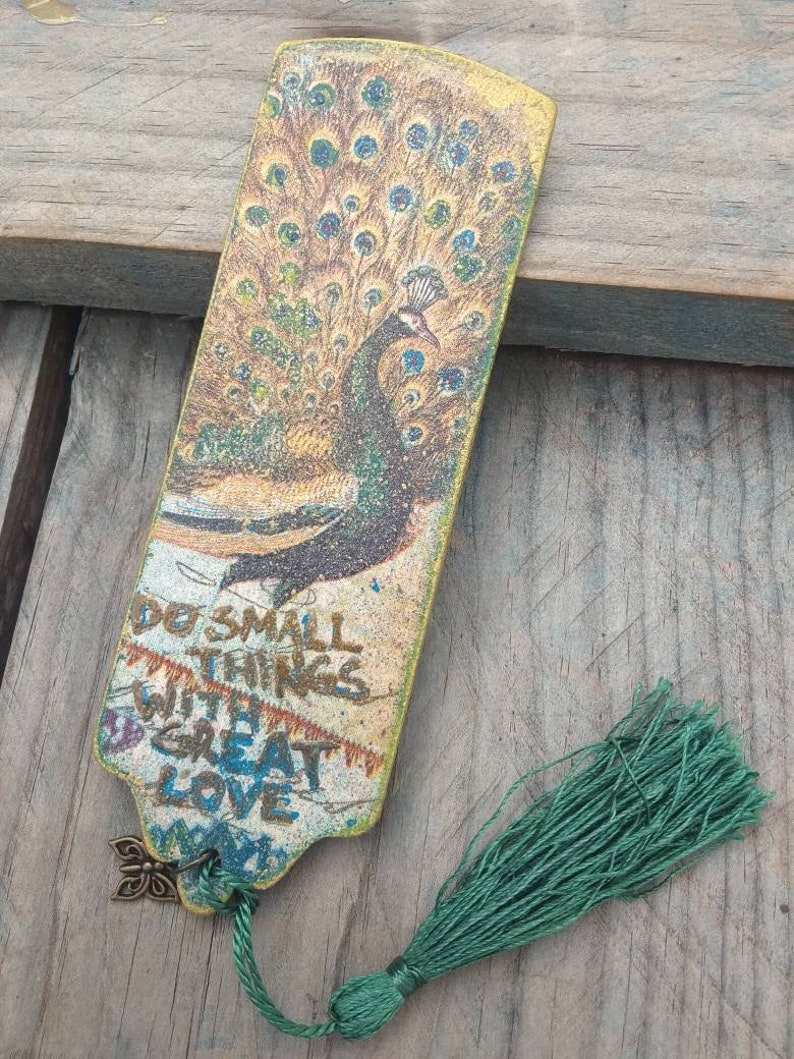 May include: A wooden bookmark with a peacock design and a green tassel. The bookmark has the text "Do small things with great love" printed on it.