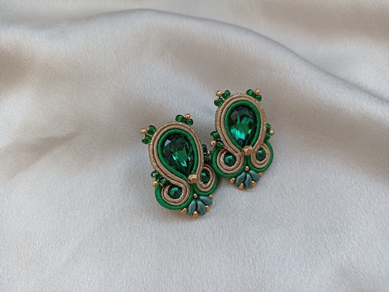 May include: A pair of green and gold earrings with a teardrop-shaped green gemstone in the center of each earring.