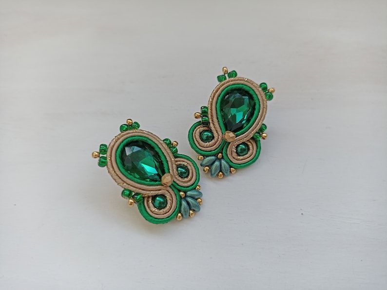 May include: A pair of green and gold earrings with a teardrop-shaped green gemstone in the center of each earring.
