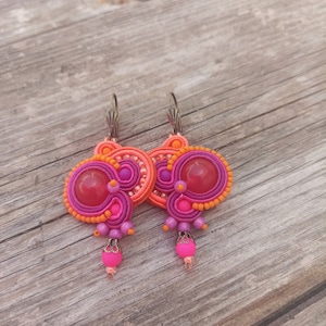 Puede incluir: Un par de pendientes de color rosa y naranja con una piedra rosa grande en el centro de cada pendiente. Los pendientes están hechos de arcilla polimérica y tienen un diseño delicado e intrincado.