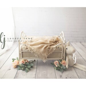 May include: A white metal crib with a beige blanket and a white unicorn stuffed animal. The crib is on a white wooden floor with a floral arrangement in front of it.