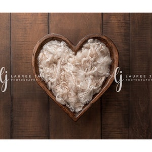 May include: A heart-shaped wooden bowl filled with fluffy white faux fur, perfect for newborn photography props. The bowl is on a rustic brown wooden background.