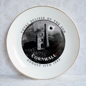 May include: A white ceramic plate with a gold rim features a black and white image of a building with a chimney and the text "Total Eclipse of the Sun Cornwall August 11th 1999".