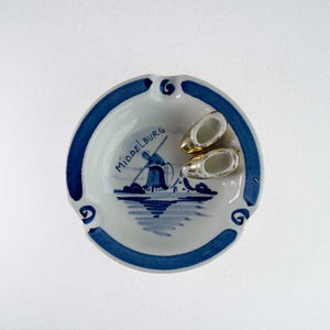 Może przedstawiać: Okrągła, biała ceramiczna popielniczka z niebieską obwódką i niebiesko-białą ilustracją wiatraka oraz napisem "Middelburg". Wewnątrz popielniczki znajdują się dwa miniaturowe chodaki ze złotymi i srebrnymi akcentami.