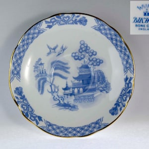 Pode incluir: Um pires de porcelana fina Duchess com padrão Willow e borda dourada. O pires apresenta uma cena de paisagem azul com pagodes, árvores e pássaros. A borda tem um design floral e geométrico azul. O logotipo Duchess Bone China England é visível.
