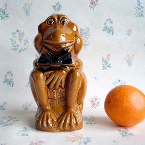 Peut inclure: Une figurine de grenouille en céramique brune portant un nœud papillon noir. La grenouille est assise les jambes croisées et porte une étiquette avec le nom "Fred".