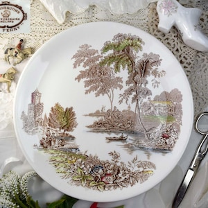 May include: White ceramic plate with a brown and green landscape scene. The plate features a river, trees, and a building. The plate is marked "Swinnerton's Staffordshire England" and "The Ferry".