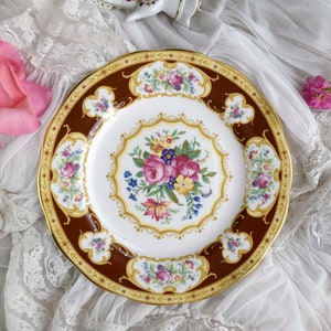 Op de afbeelding: Een decoratief bord met een bruine rand en een gouden rand. Het bord heeft een centraal bloemmotief met roze, paarse en gele bloemen. Het bord heeft een wit midden en is omgeven door een bruine rand met bloemaccenten.