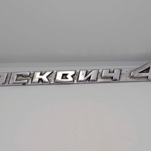 Puede incluir: Un emblema metálico plateado con la palabra cirílica "Москвич" y el número "412". El emblema rectangular tiene una apariencia ligeramente desgastada, lo que sugiere que es vintage.